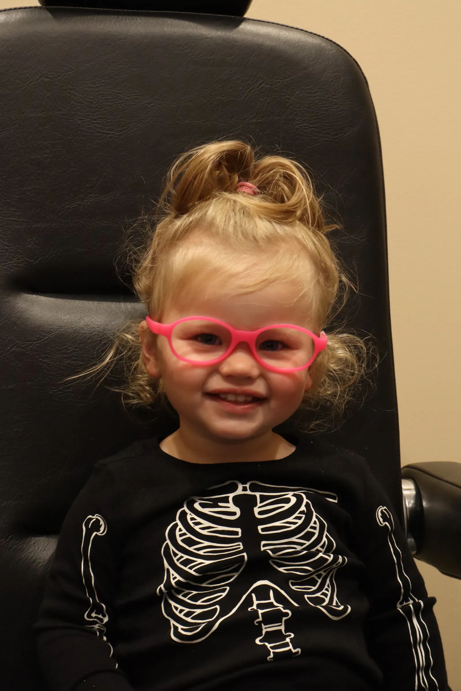 A smiling young child wearing pink eyeglasses in the exam chair at West & Stannish